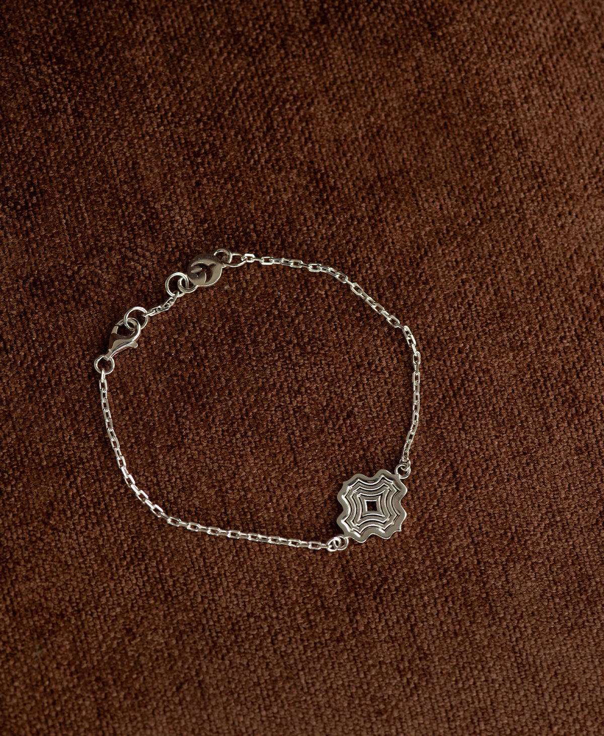 The Alma Bracelet featuring a silver pendant with a concentric wave design, on a textured brown fabric background.