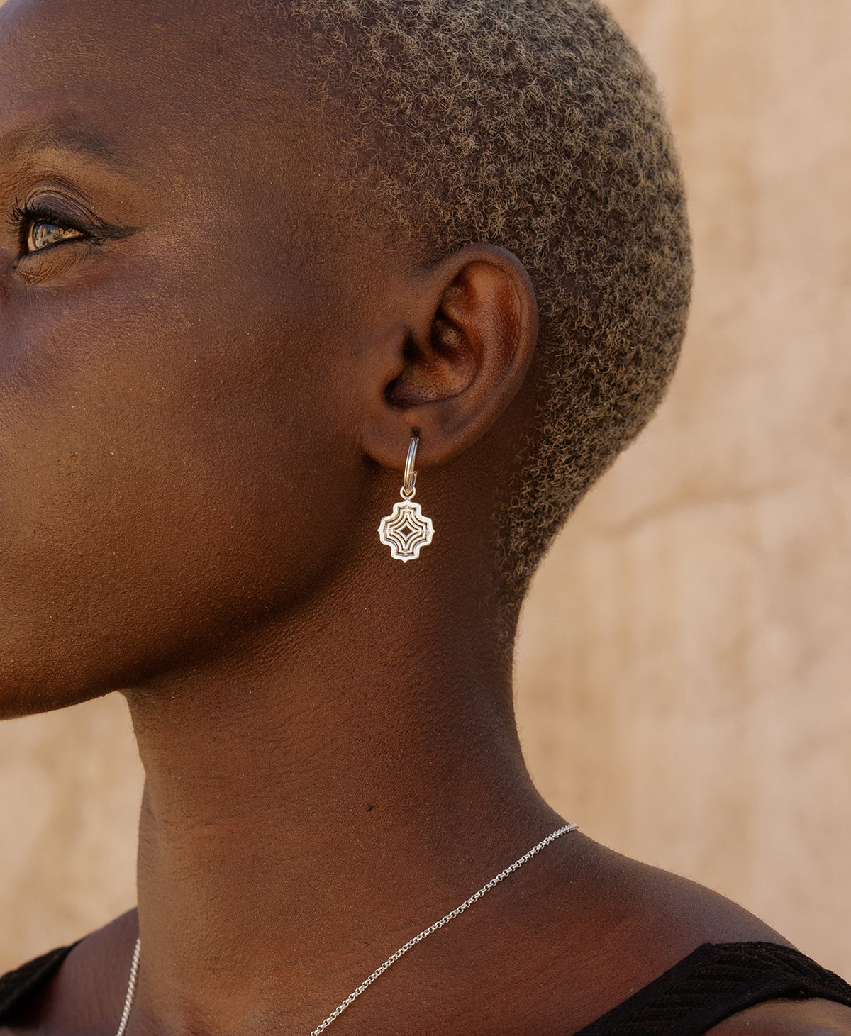A close-up side profile of a person wearing the Alma Hoops, featuring a silver pendant with a concentric wave design, suspended from a hoop.