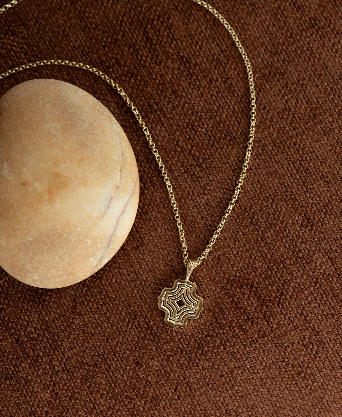 The Alma Necklace featuring a silver pendant with a concentric wave design, next to a cream stone on a textured brown fabric background.