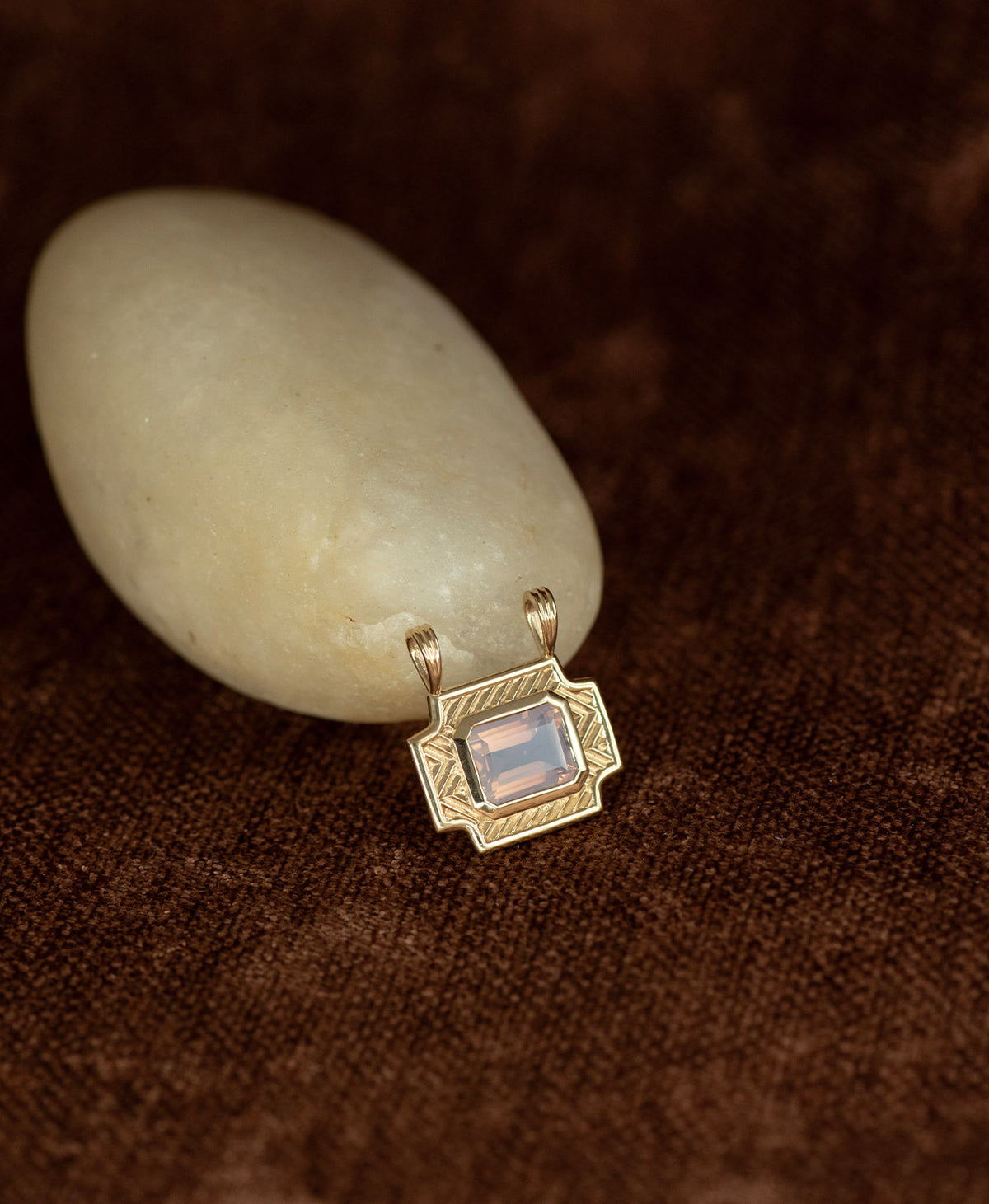 A gold pendant with a rectangular lavender quartz stone, resting against a smooth white stone on a textured brown background.