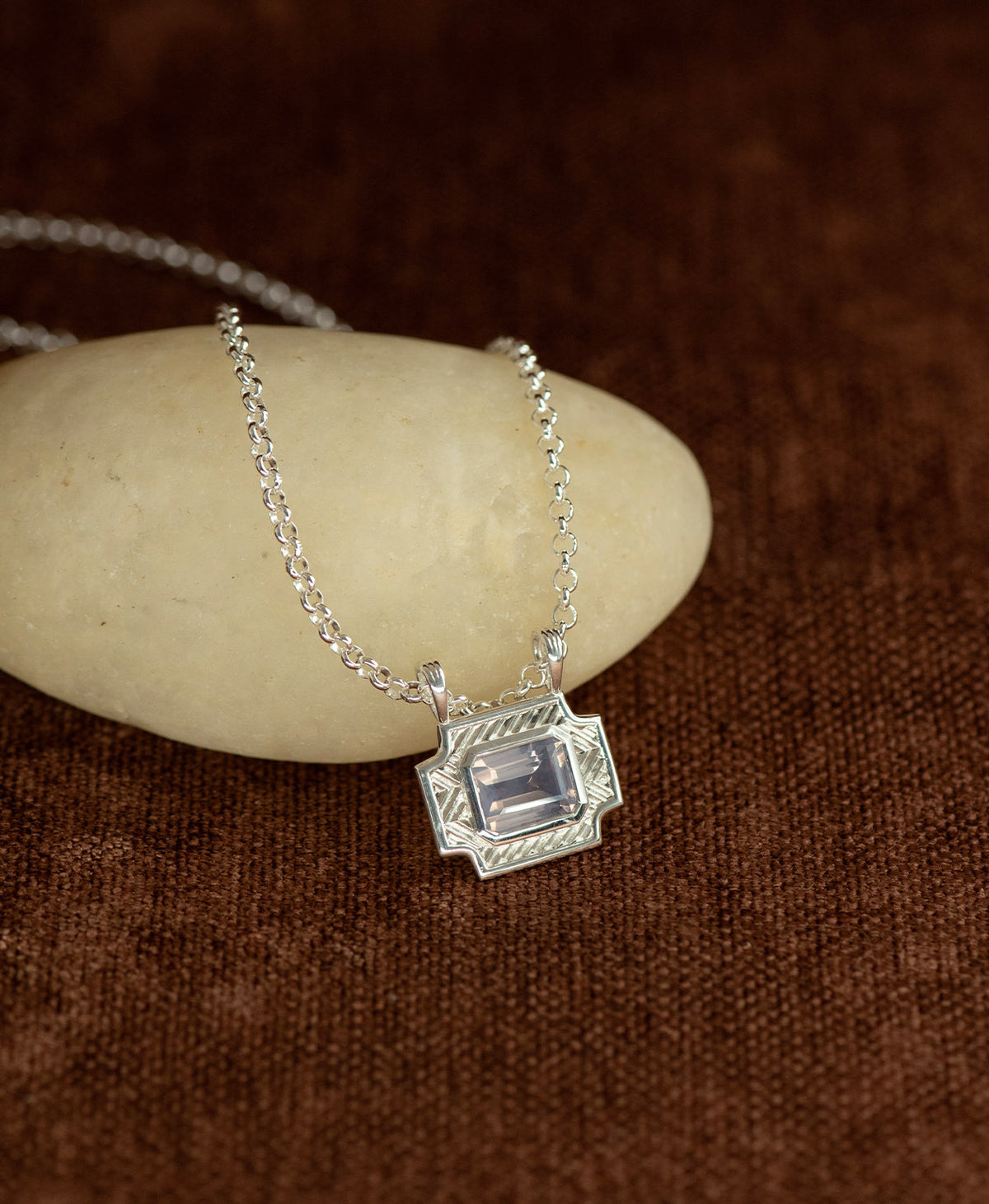 A silver pendant with a rectangular lavender quartz stone, resting on a cream stone hanging on a belcher chain against a textured brown background.