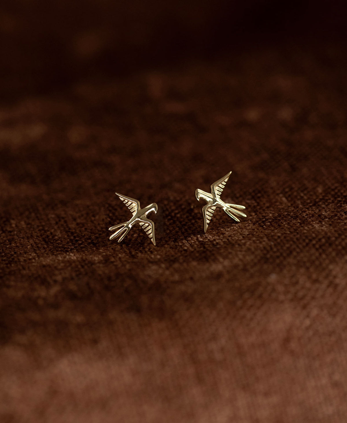 A pair of gold bird stud earrings with detailed feather and wing textures, displayed on a dark brown textured fabric background.