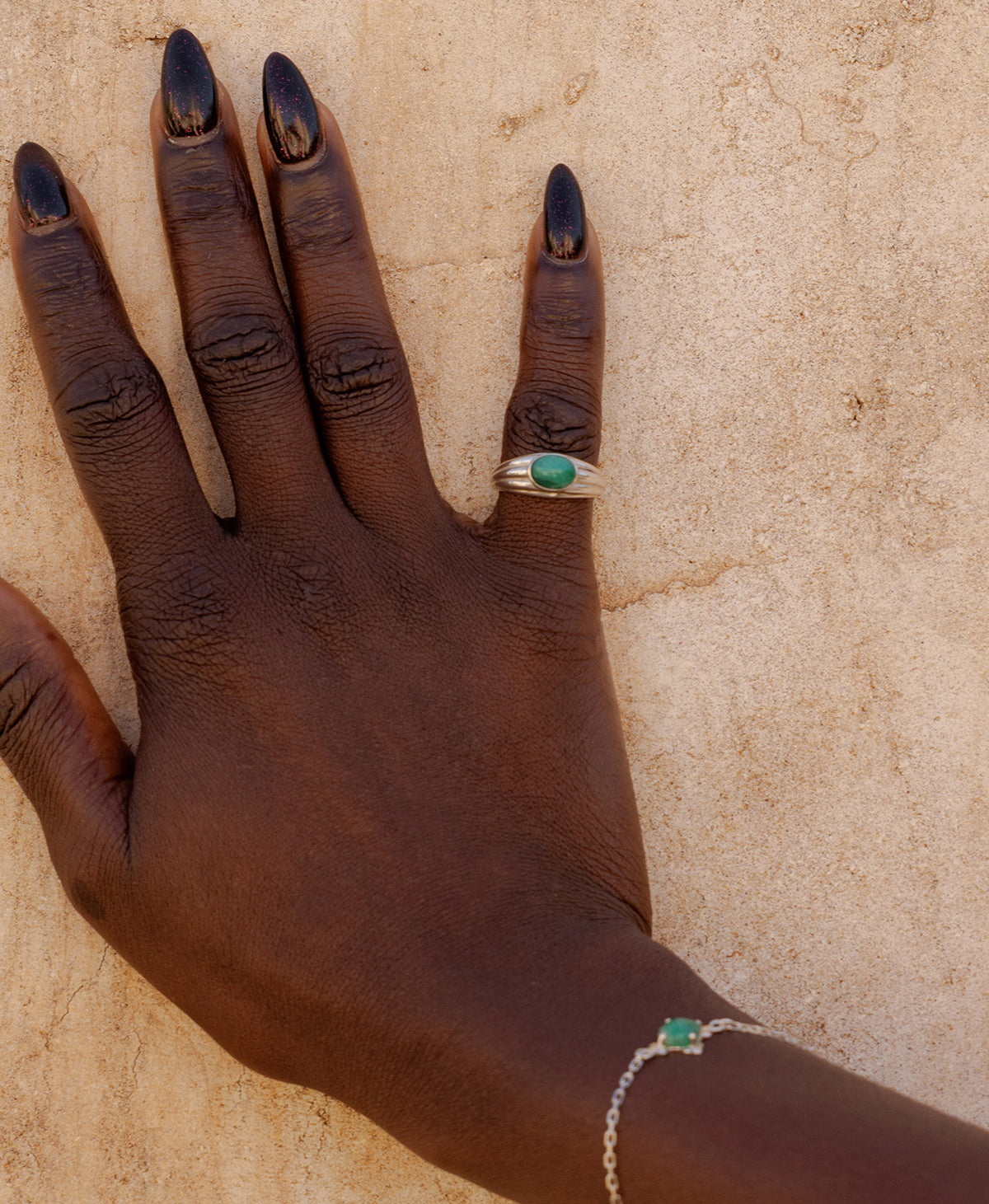 A closeup of a hand wearing the silver Esmeralda Ring, featuring a cabochon cut Emerald set in a graduated band, worn on the pinky finger and paired with a silver, emerald bracelet.