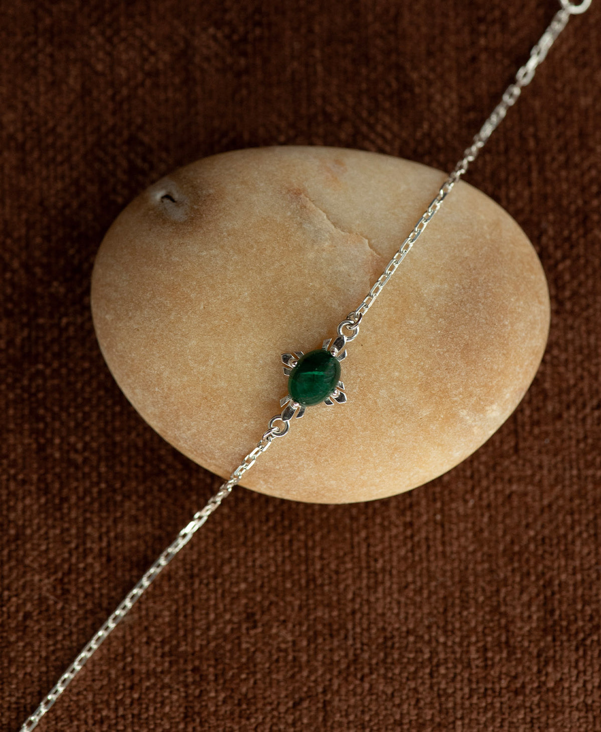 The silver Farah Bracelet featuring a cabochon cut emerald, layered over a cream stone on a textured brown fabric background.