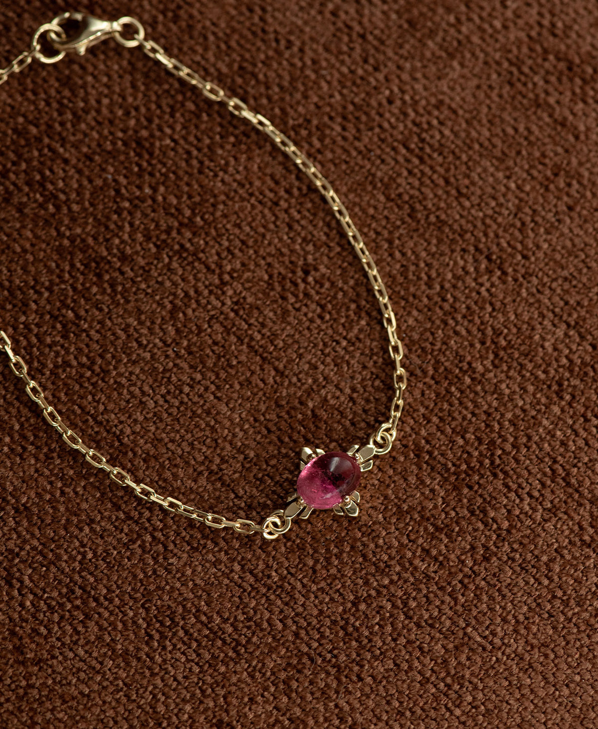 A close up of the gold Farah Bracelet featuring a cabochon cut pink tourmaline, on a textured brown fabric background.