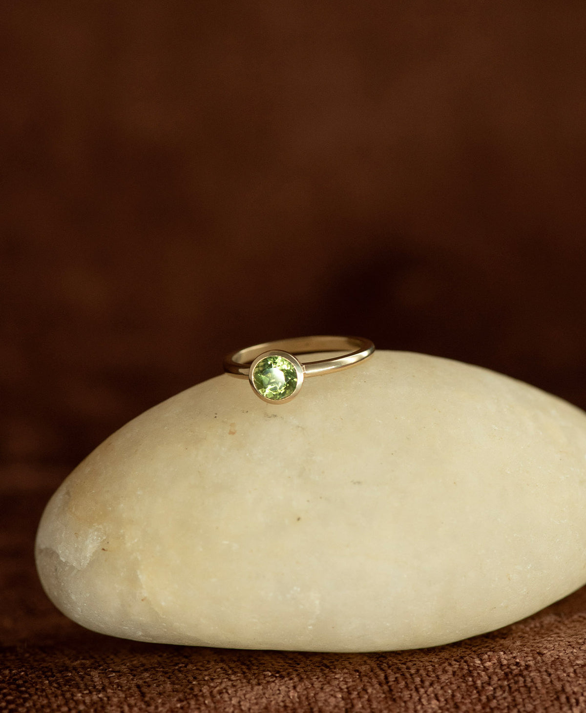 A close up of the gold Mon Petit Stacker Ring with a bezel set, round cut Peridot resting on a cream stone on a textured brown background.