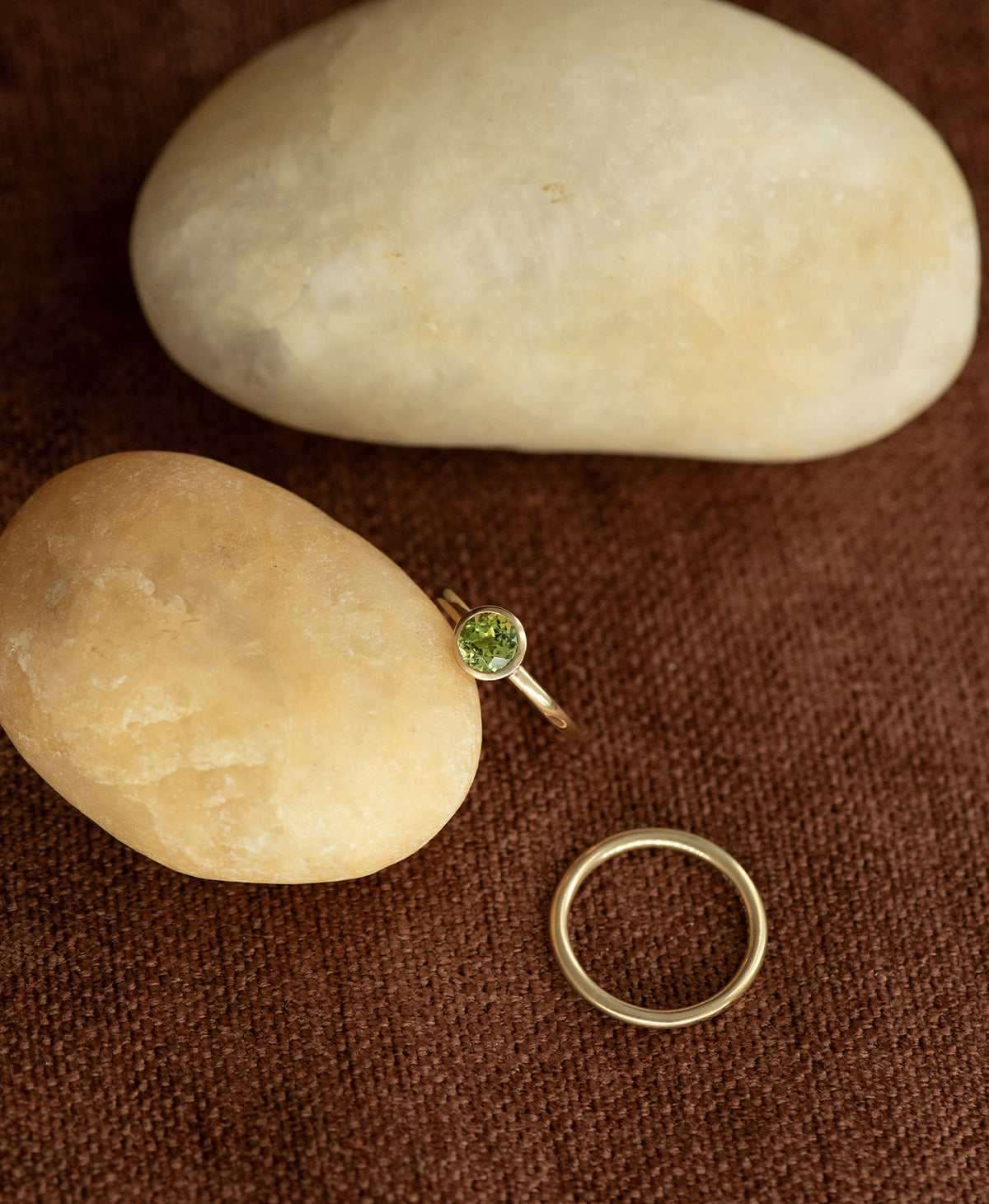 The gold Mon Petit Stacker Ring with a bezel set, round cut Peridot rests on a cream stone on a textured brown background with a plain gold band to the side.