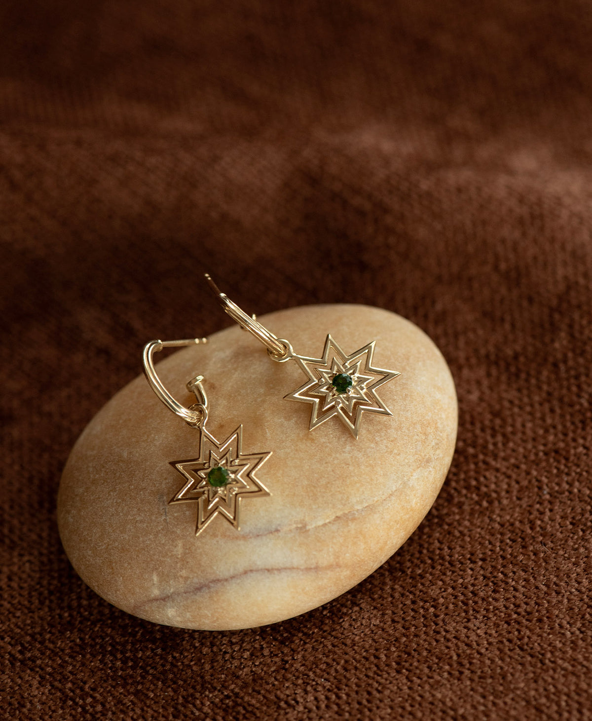 The gold Nora Hoops, featuring an eight pointed star suspended from a hoop, displayed on a smooth beige stone against a textured brown fabric background.