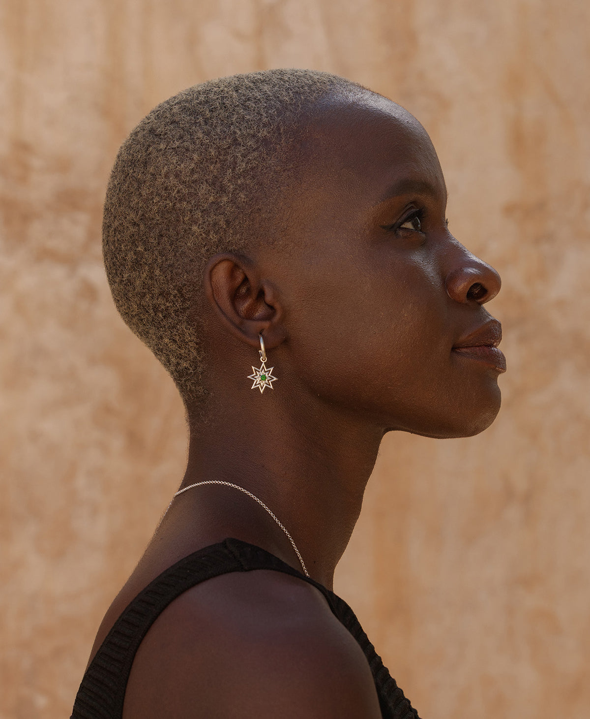 A close-up of a person wearing the silver Nora Hoops, featuring a gold star hanging from a hoop with a central green stone.