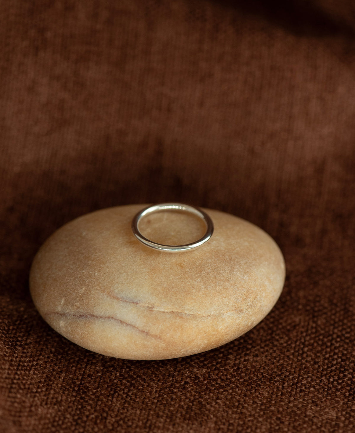 The smooth, curved Selma stacking ring in 925 sterling silver, rests on a cream stone on a textured brown background.