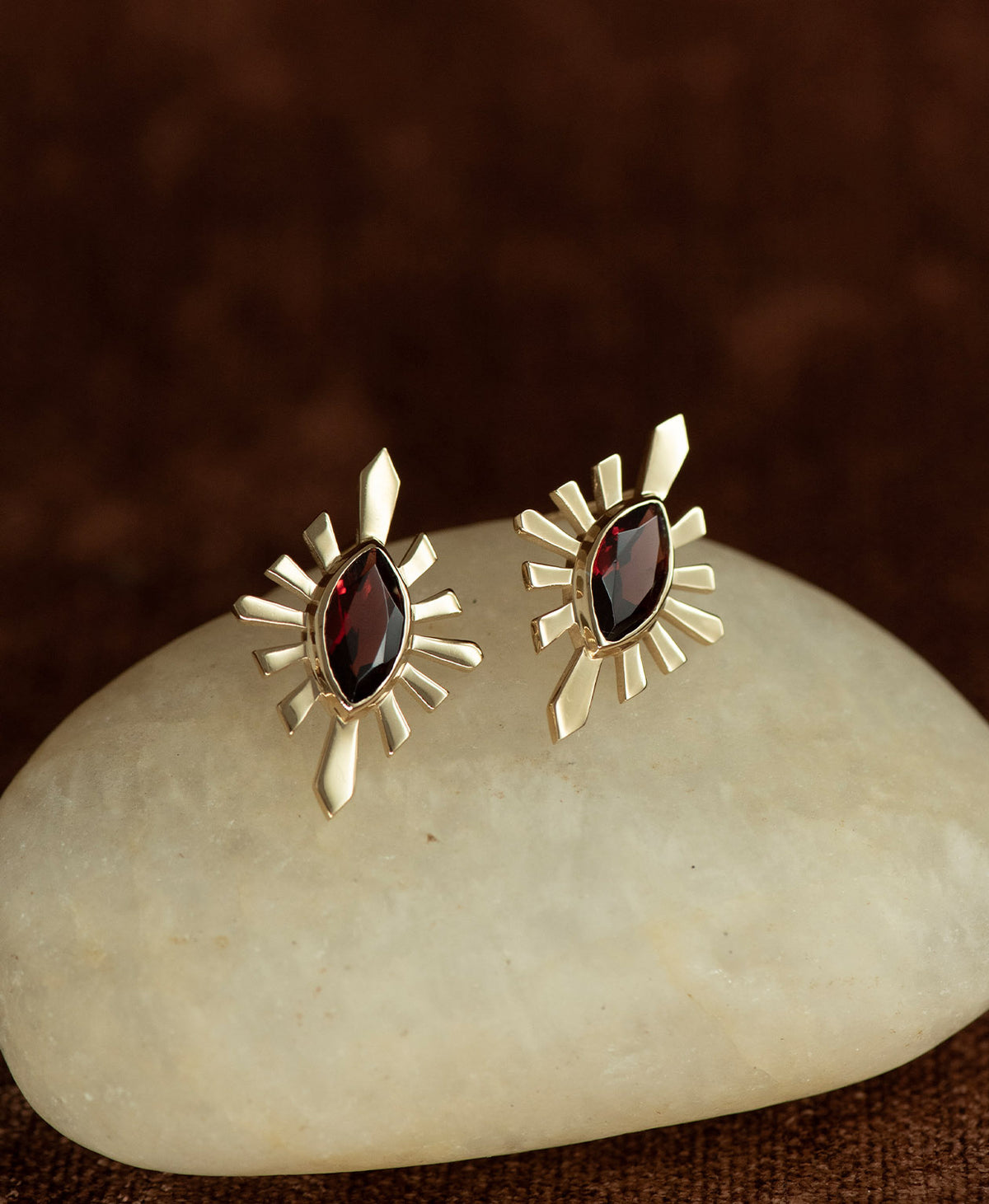 The Sister Studs, featuring a central marquise cut Garnet nestled within a gold sunburst frame, displayed on a smooth beige stone against a textured brown fabric background.