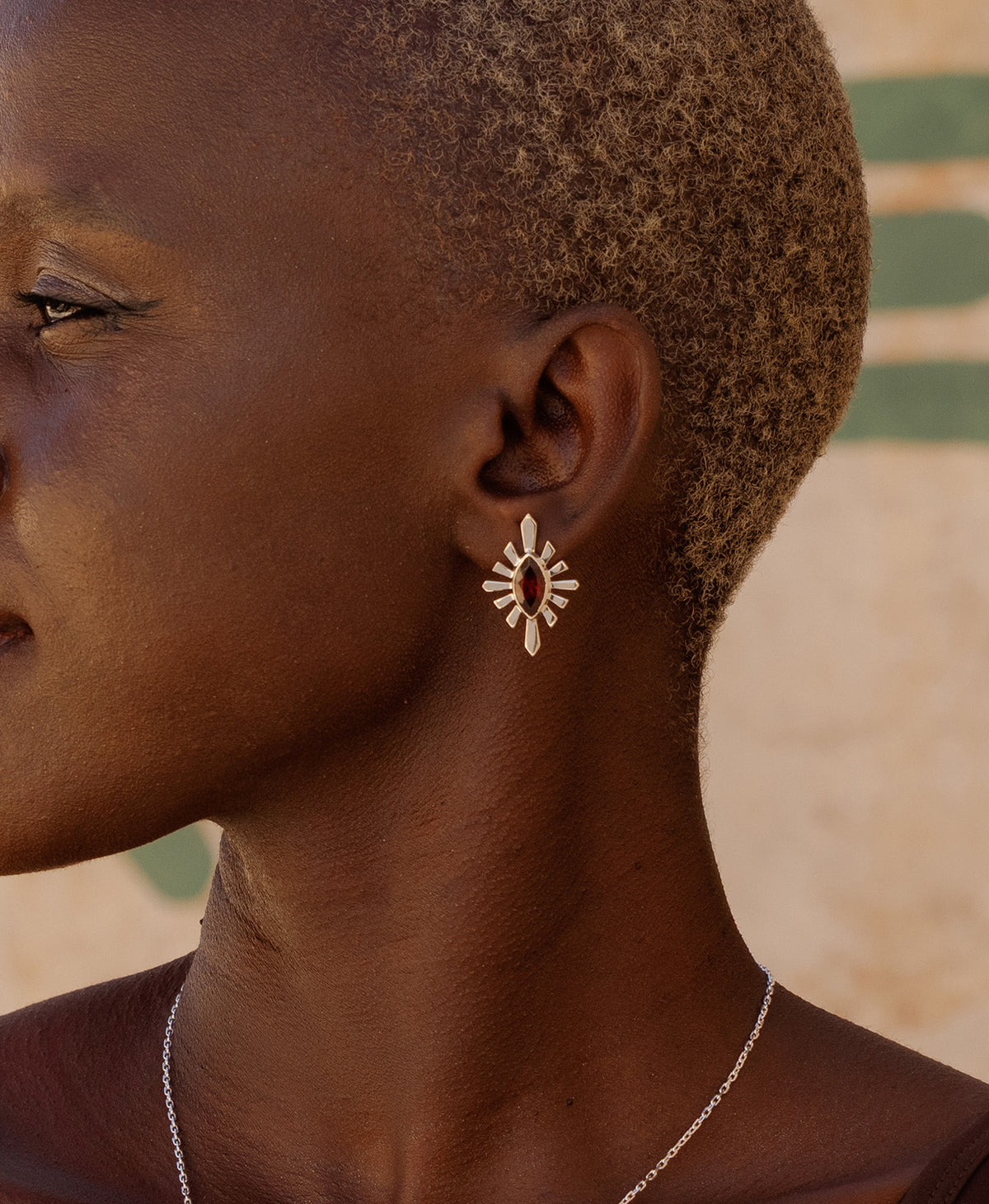 A close-up side profile of a person wearing the Sister Studs, featuring a marquise cut garnet in a silver linear sunburst frame.