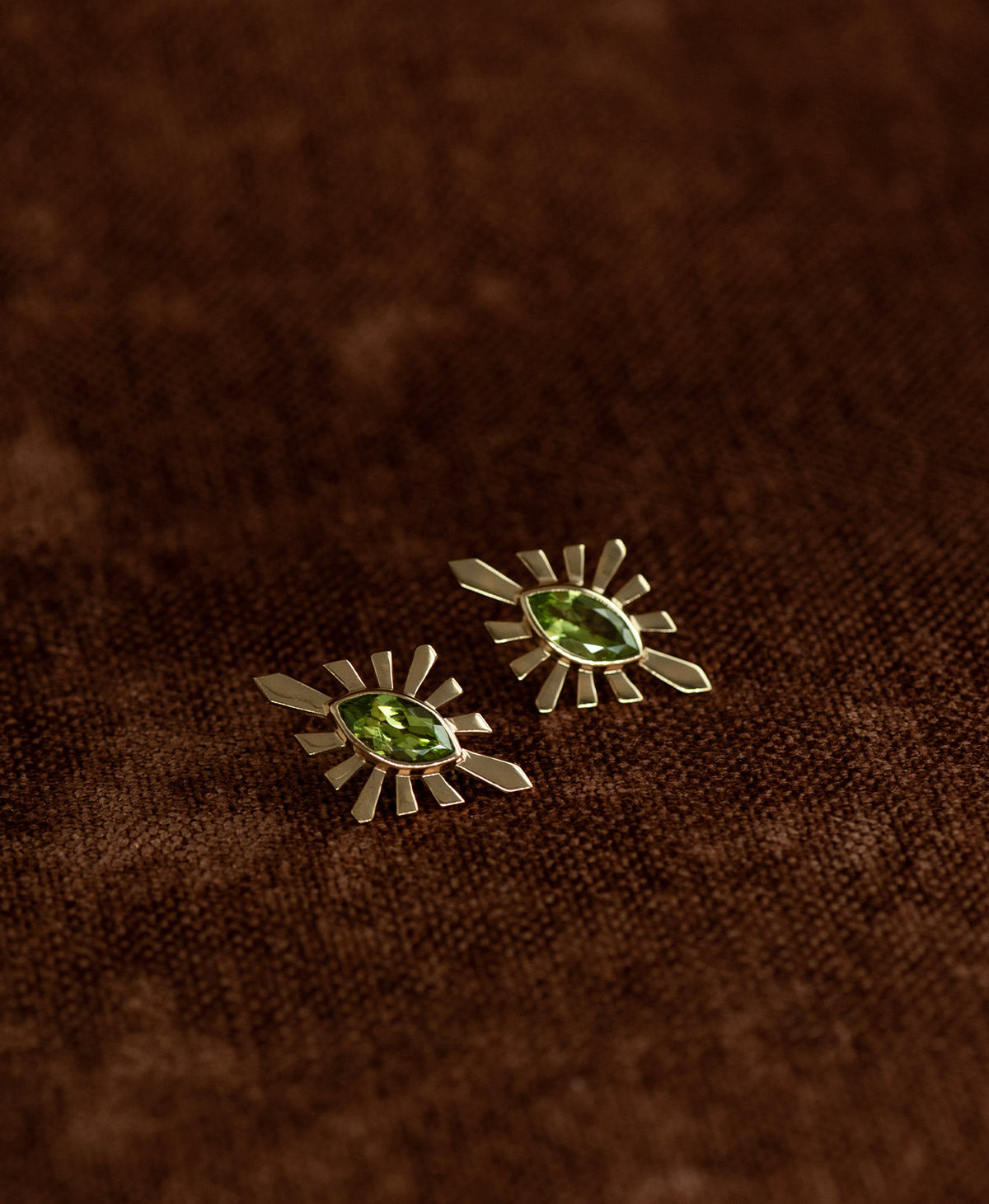 The Sister Studs, featuring a central marquise cut Peridot nestled within a silver sunburst frame, displayed against a textured brown fabric background.