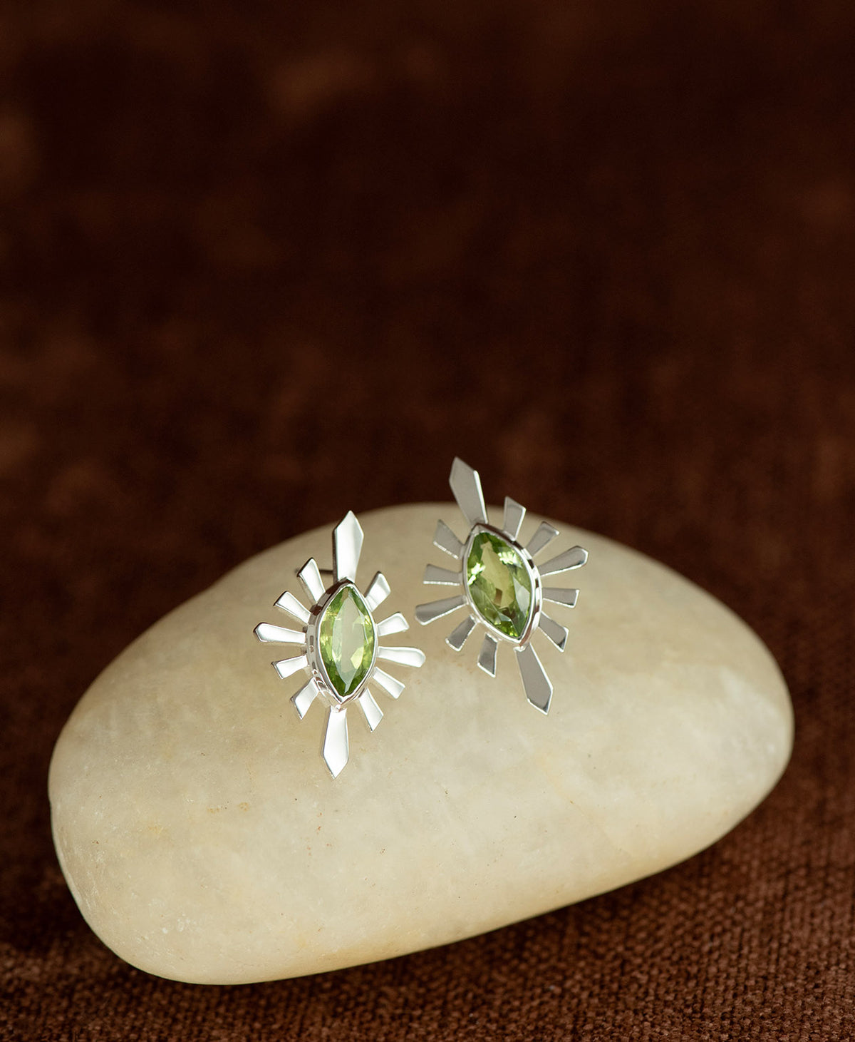 The Sister Studs, featuring a central marquise cut Peridot nestled within a silver sunburst frame, displayed on a smooth beige stone against a textured brown fabric background.