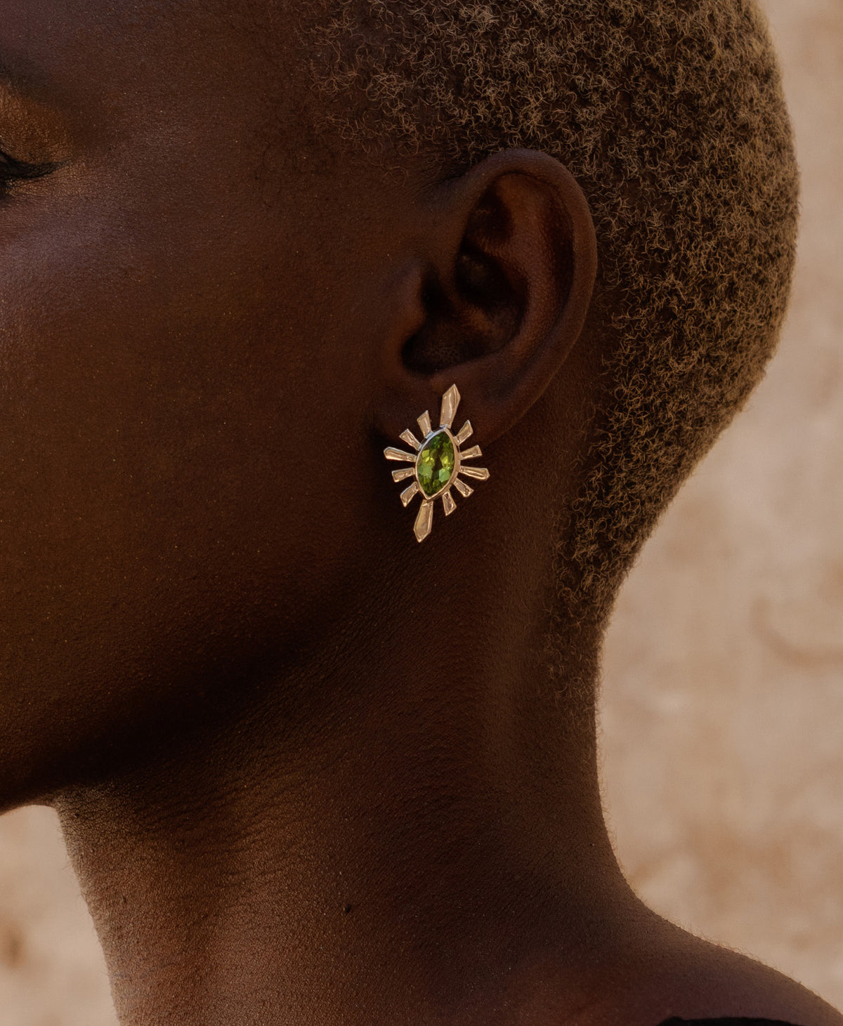 A close-up side profile of a person wearing the Sister Studs, featuring a marquise cut peridot in a silver linear sunburst frame.