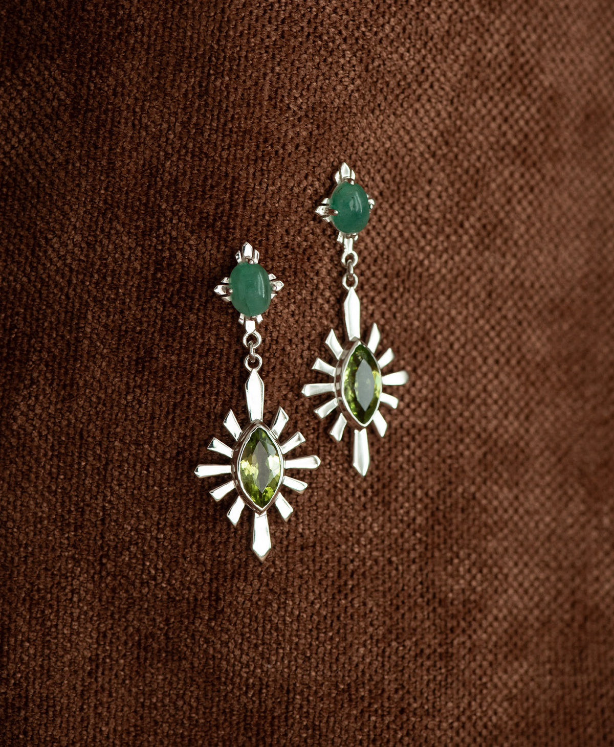 A pair of silver Sisterhood Earrings with a cabochon cut Emerald, set within a linear border, paired with a marquise cut Peridot nestled with a sunburst frame, hanging against a textured brown fabric background.