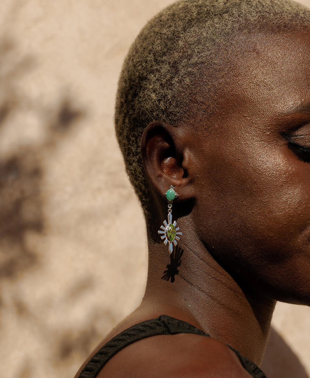 A close-up side profile of a person wearing the silver Sisterhood Earring, featuring a cabochon cut emerald set within a linear border, paired with a marquise cut peridot with a sunburst frame.