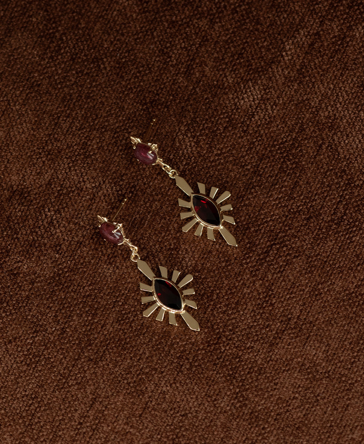 A pair of gold Sisterhood Earrings with a cabochon cut pink Tourmaline, set within a linear border, paired with a marquise cut Garnet nestled with a sunburst frame, displayed against a textured brown fabric background.