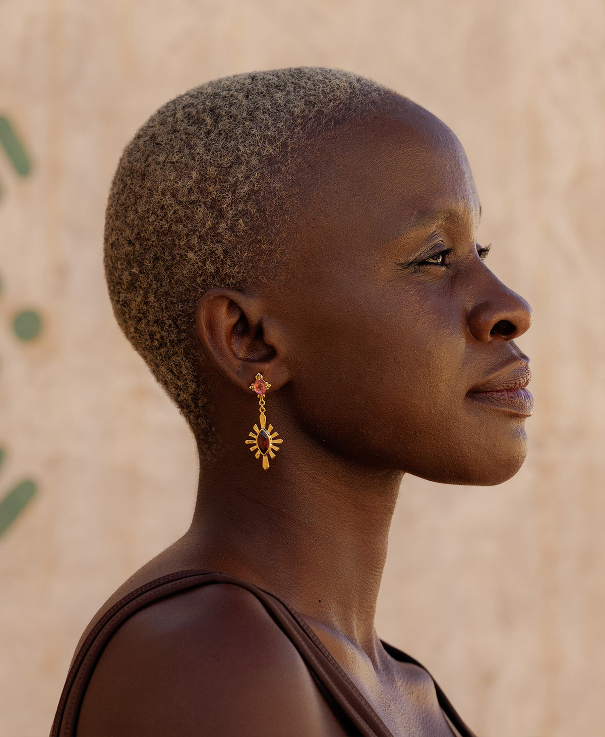 A side profile of a person wearing the Sisterhood Earring, featuring a cabochon cut pink Tourmaline is set within a linear border, paired with a marquise cut Garnet with a sunburst frame.
