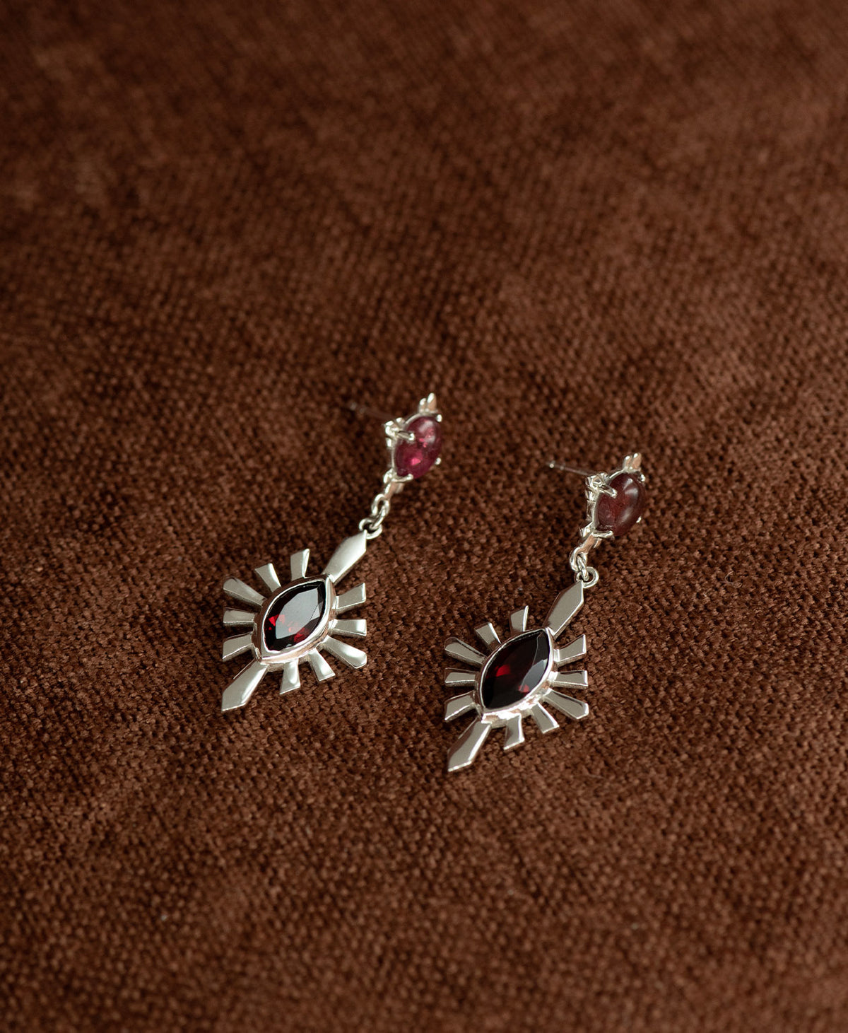 A pair of silver Sisterhood Earrings with a cabochon cut pink Tourmaline, set within a linear border, paired with a marquise cut Garnet nestled with a sunburst frame, displayed against a textured brown fabric background.