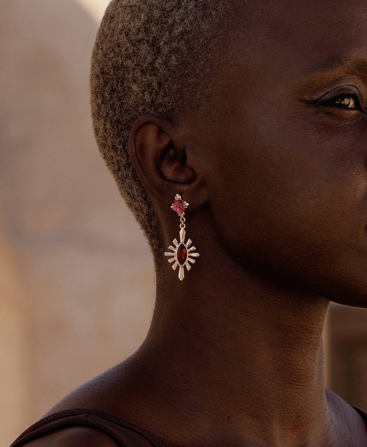 A close-up side profile of a person wearing the silver Sisterhood Earring, featuring a cabochon cut pink Tourmaline is set within a linear border, paired with a marquise cut Garnet with a sunburst frame.