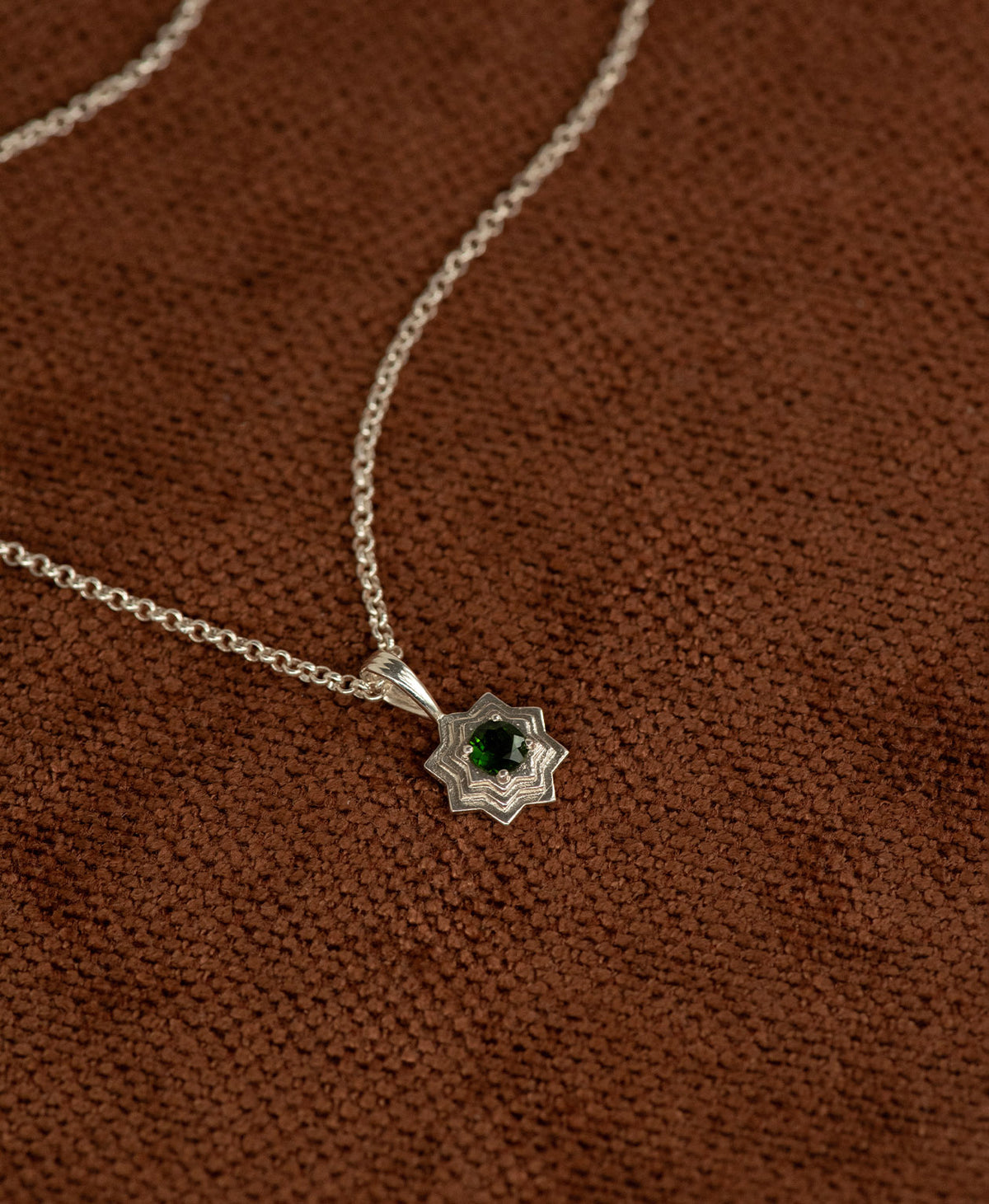 Close up of the Sitara Necklace featuring a silver star like pendant with concentric lines radiating outwards from a Chrome Diopside resting at its centre, displayed on a textured brown fabric background.