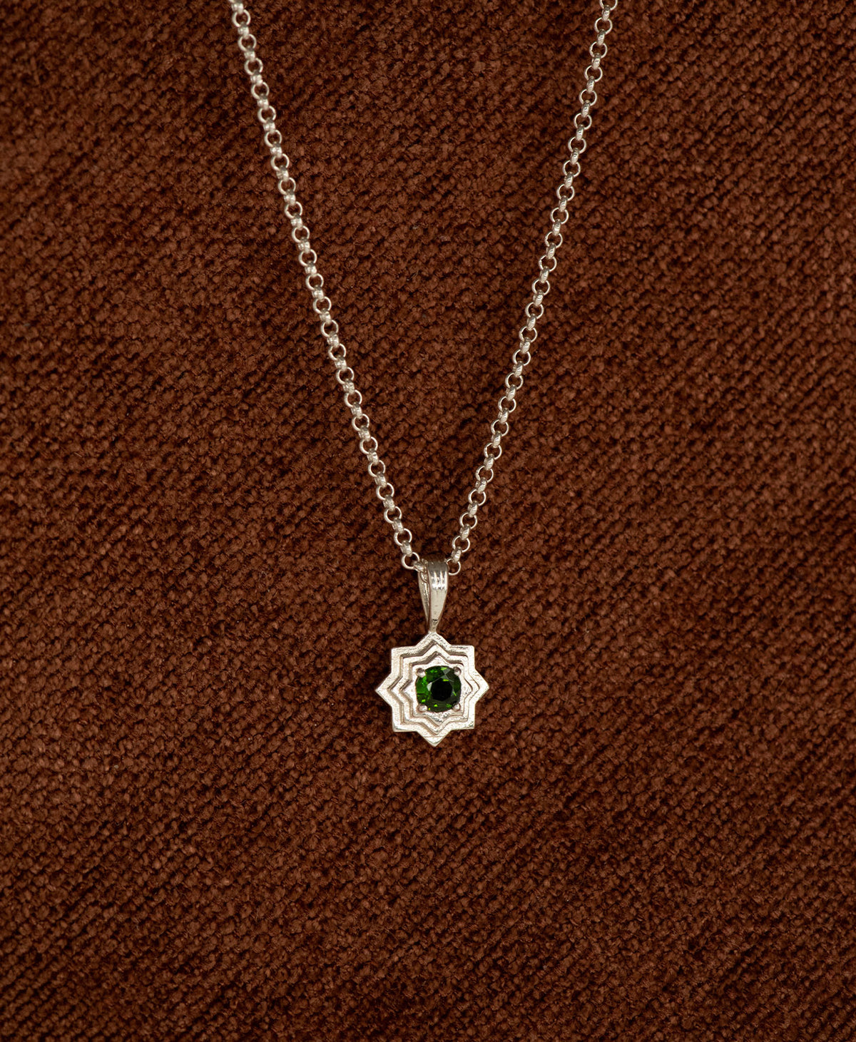 The Sitara Necklace featuring a silver star like pendant with concentric lines radiating outwards from a Chrome Diopside resting at its centre, displayed on a textured brown fabric background.