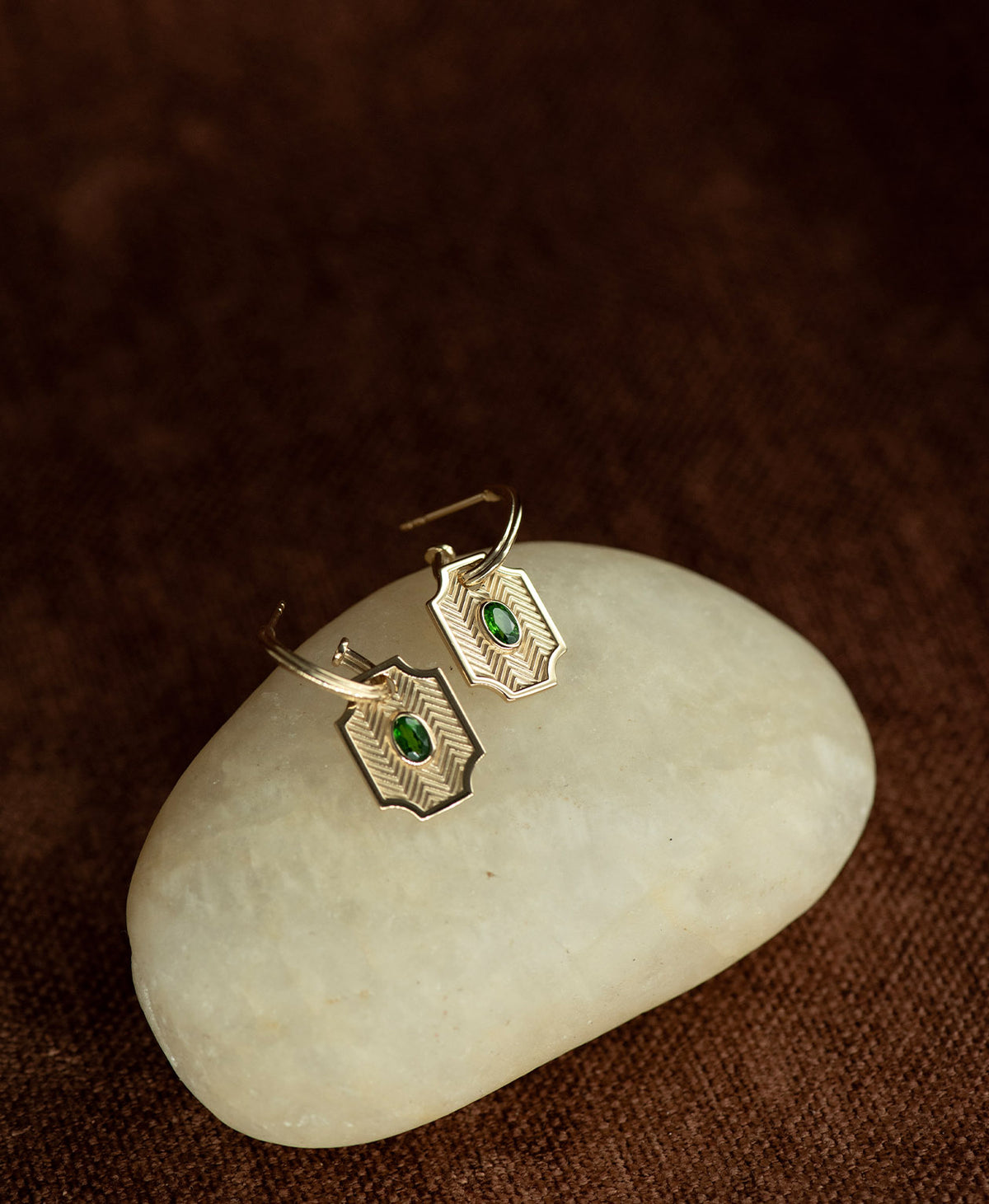 The Soul Sister Hoops, featuring a gold geometric pendant threaded onto a hoop, displayed on a smooth beige stone against a textured brown fabric background.