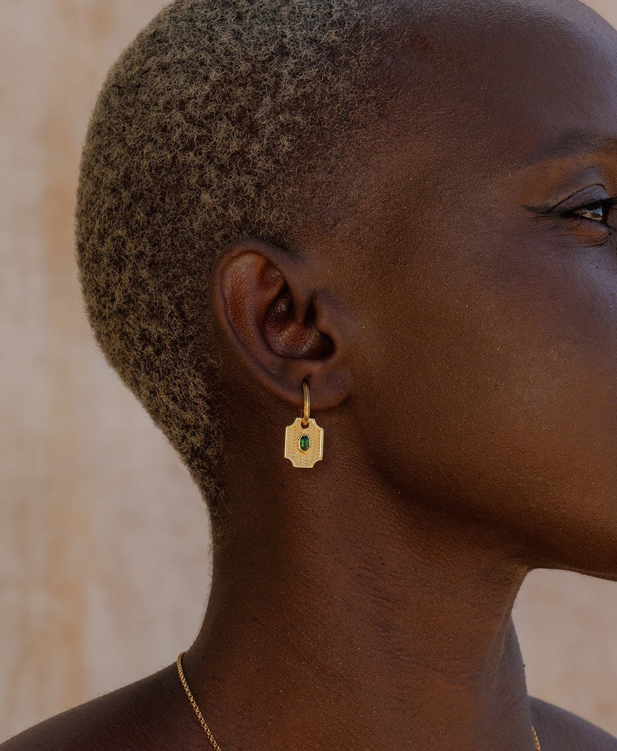 A close-up side profile of a person wearing the Soul Sister Hoops, featuring a textured gold pendant with a central green stone on a hoop.