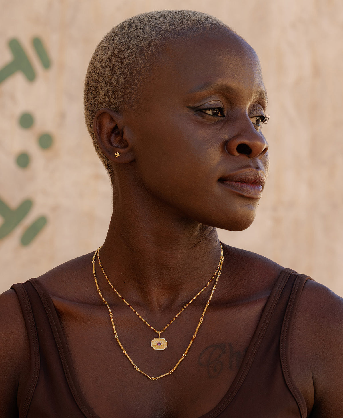 A person wearing the Soul Sister Necklace, a gold textured pendant with a central pink stone, suspended from a fine belcher chain, paired with longer chain necklace and small gold studs.