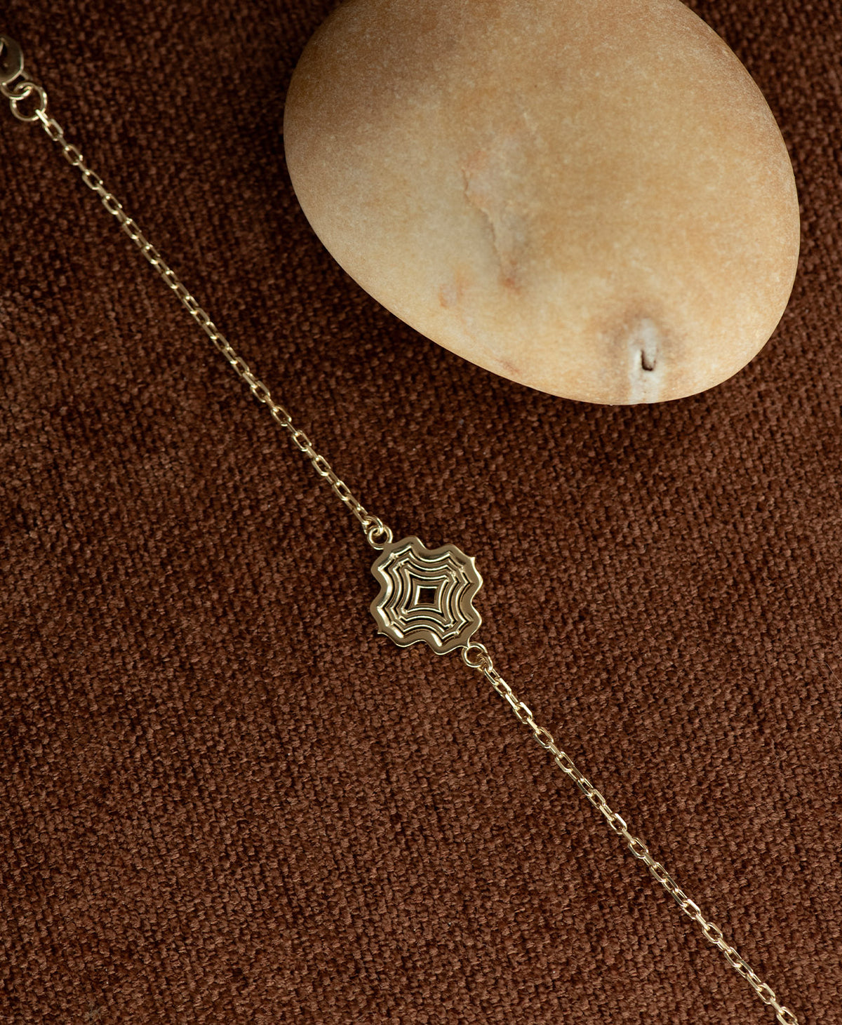 The Alma Bracelet featuring a gold pendant with a concentric wave design, resting next to a cream stone on a textured brown fabric background.