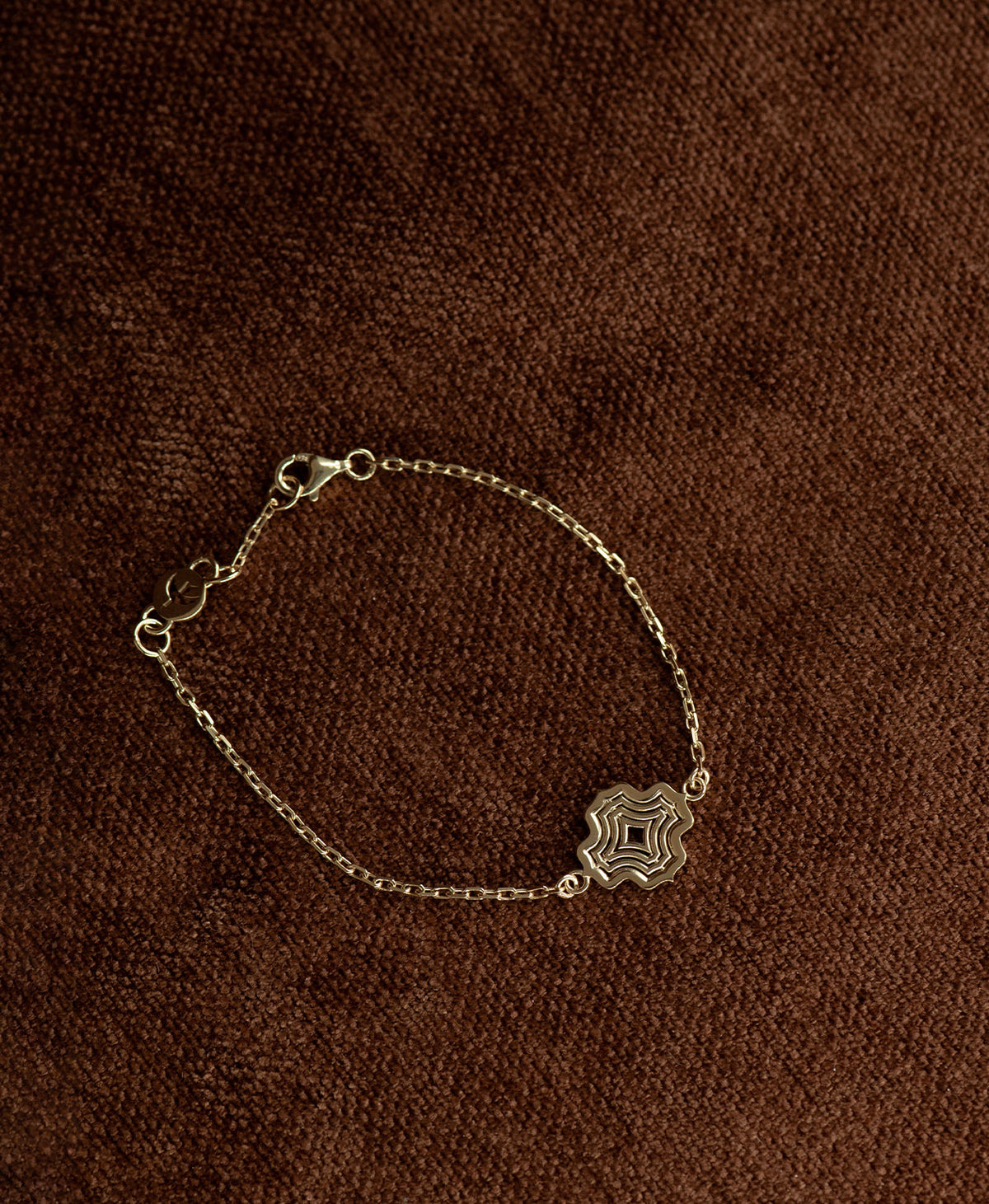 The Alma Bracelet featuring a gold pendant with a concentric wave design, on a textured brown fabric background.