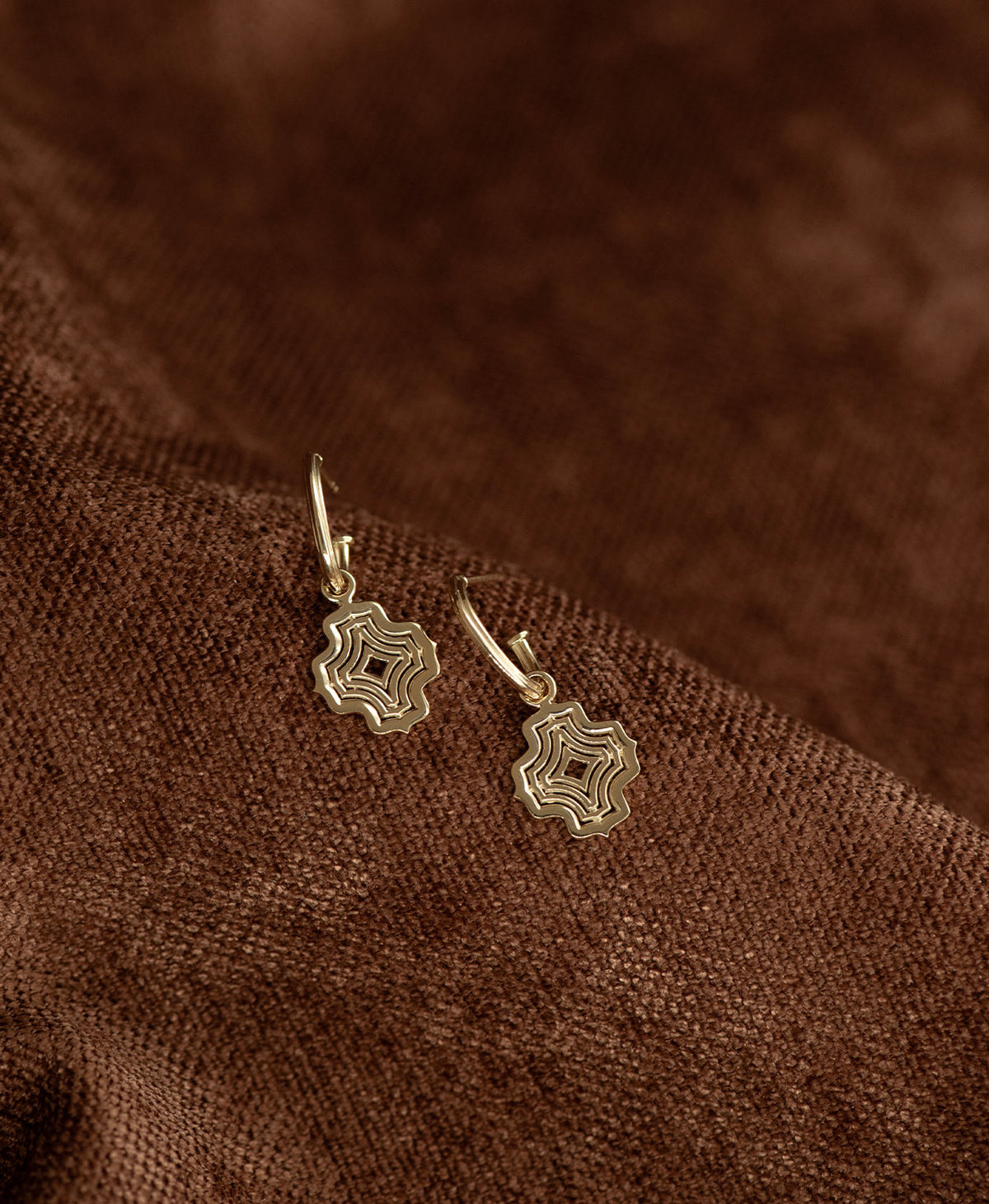 The Alma Hoops, featuring a gold pendant with a concentric wave design suspended from a hoop, displayed against a textured brown fabric background.