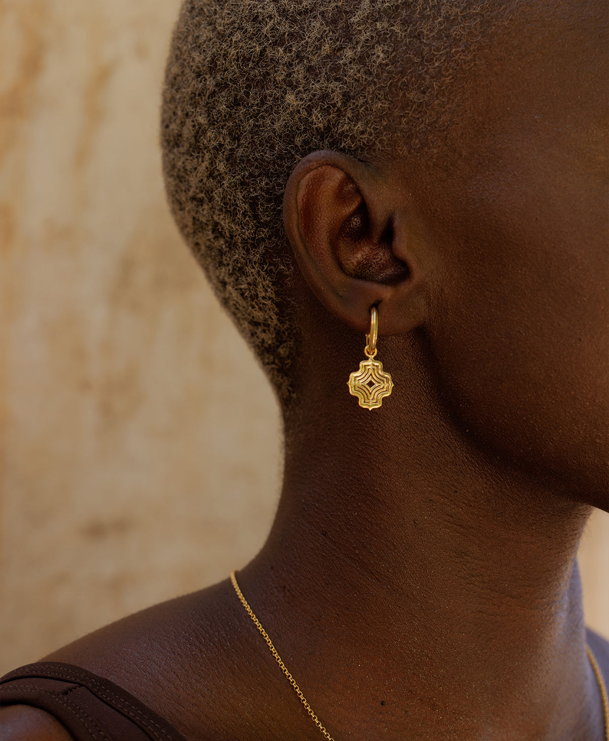 A close-up side profile of a person wearing the Alma Hoops, featuring a gold pendant with a concentric wave design, suspended from a hoop.
