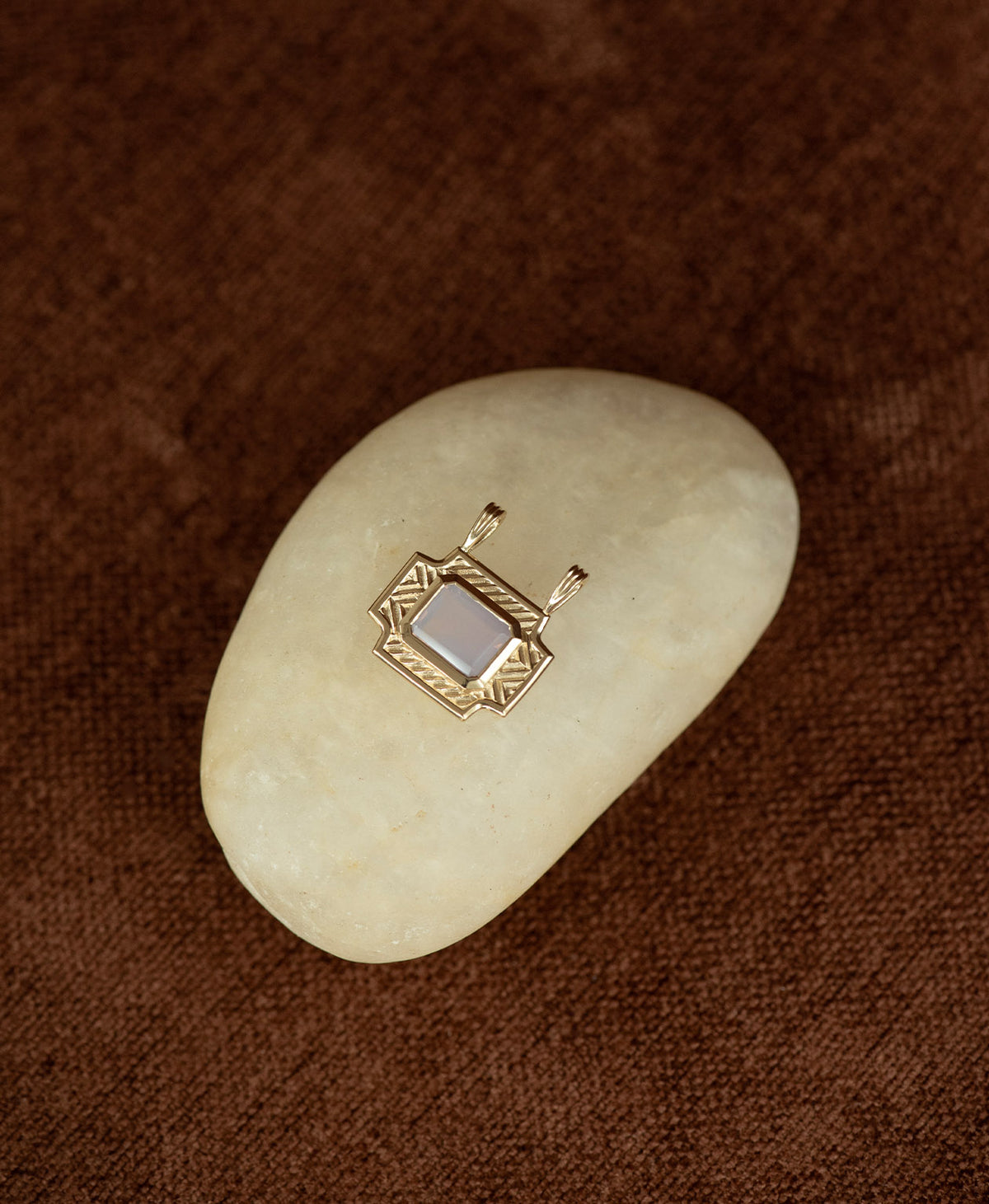 A gold pendant with a rectangular lavender quartz stone, on a smooth white stone on a textured brown background.