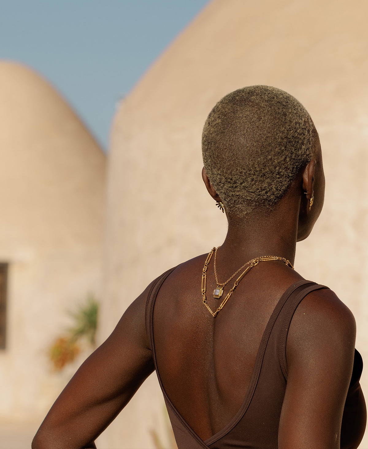 A person wearing the Ayla charm, a gold textured rectangular pendant with a central lavender stone, paired with a belcher chain, layered with a thick textured chain, resting down their back.