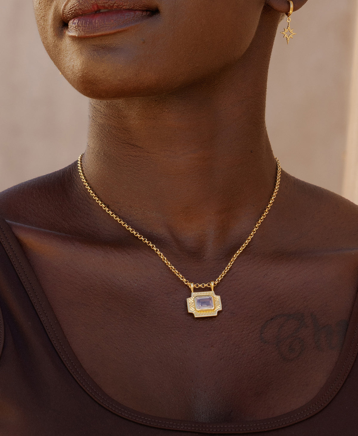 A close-up of a neck wearing the Ayla charm, a gold textured rectangular pendant with a central lavender stone, paired with a belcher chain.