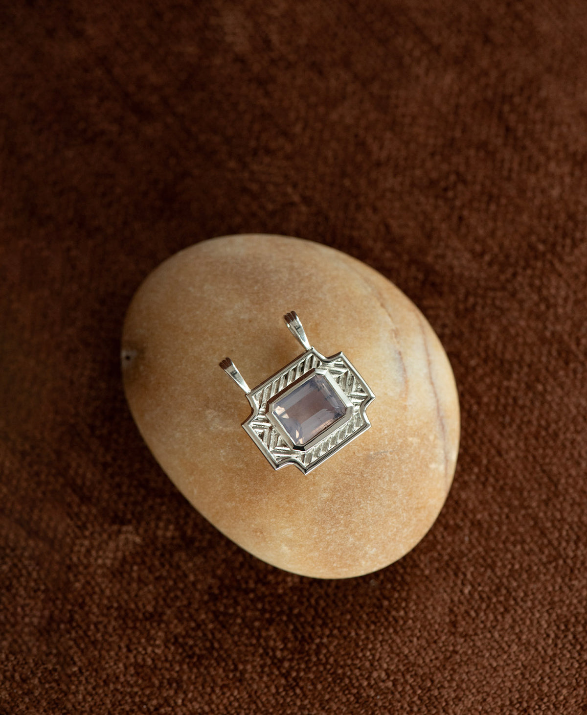 A silver pendant with a rectangular lavender quartz stone, on a smooth white stone on a textured brown background.