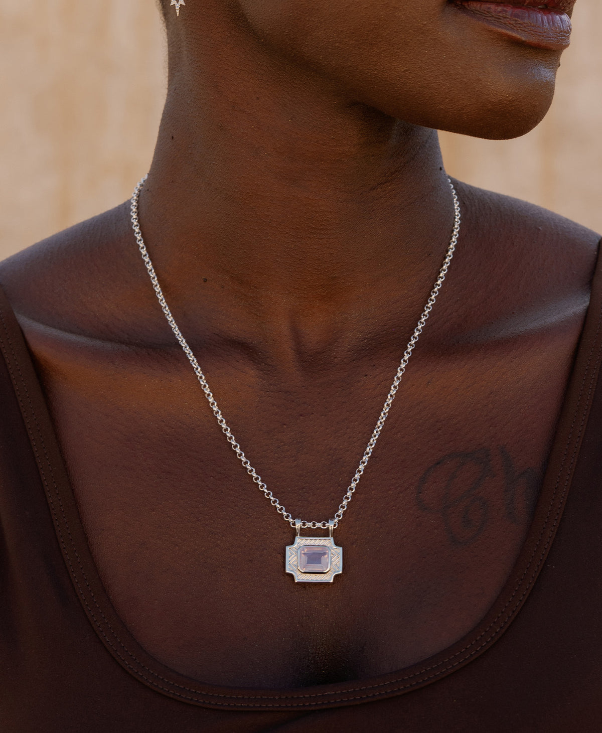 A close-up of a neck wearing the Ayla charm, a silver textured rectangular pendant with a central lavender stone, paired with a belcher chain.