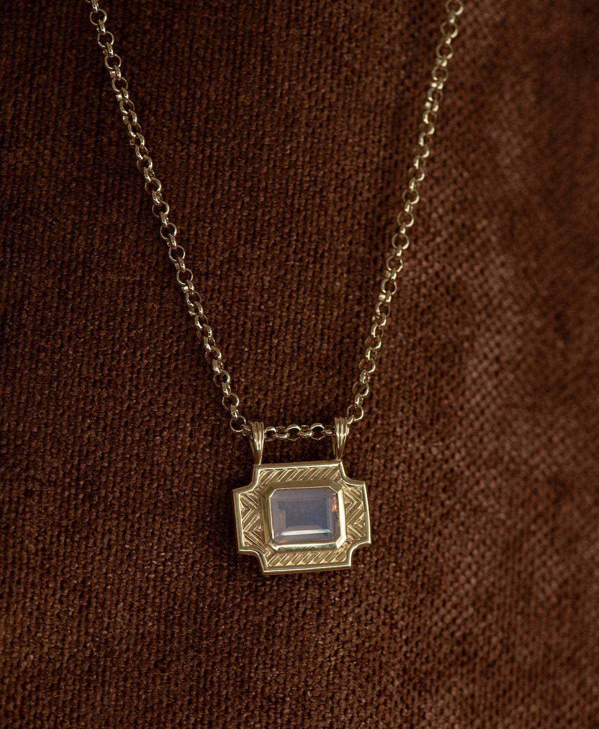 A gold pendant with a rectangular lavender quartz stone, handing on a belcher chain against a textured brown background.