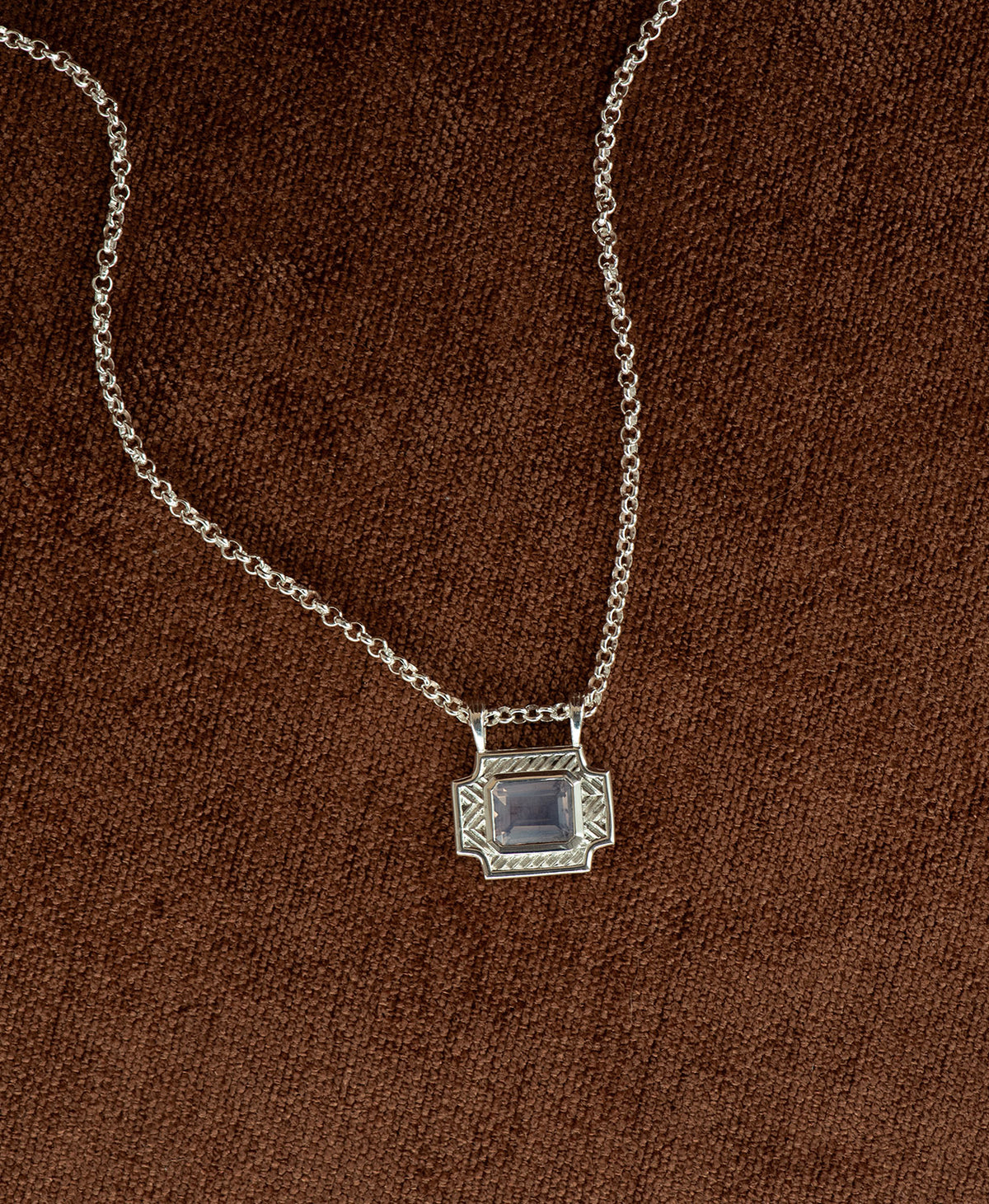 A silver pendant with a rectangular lavender quartz stone, hanging on a belcher chain against a textured brown background.