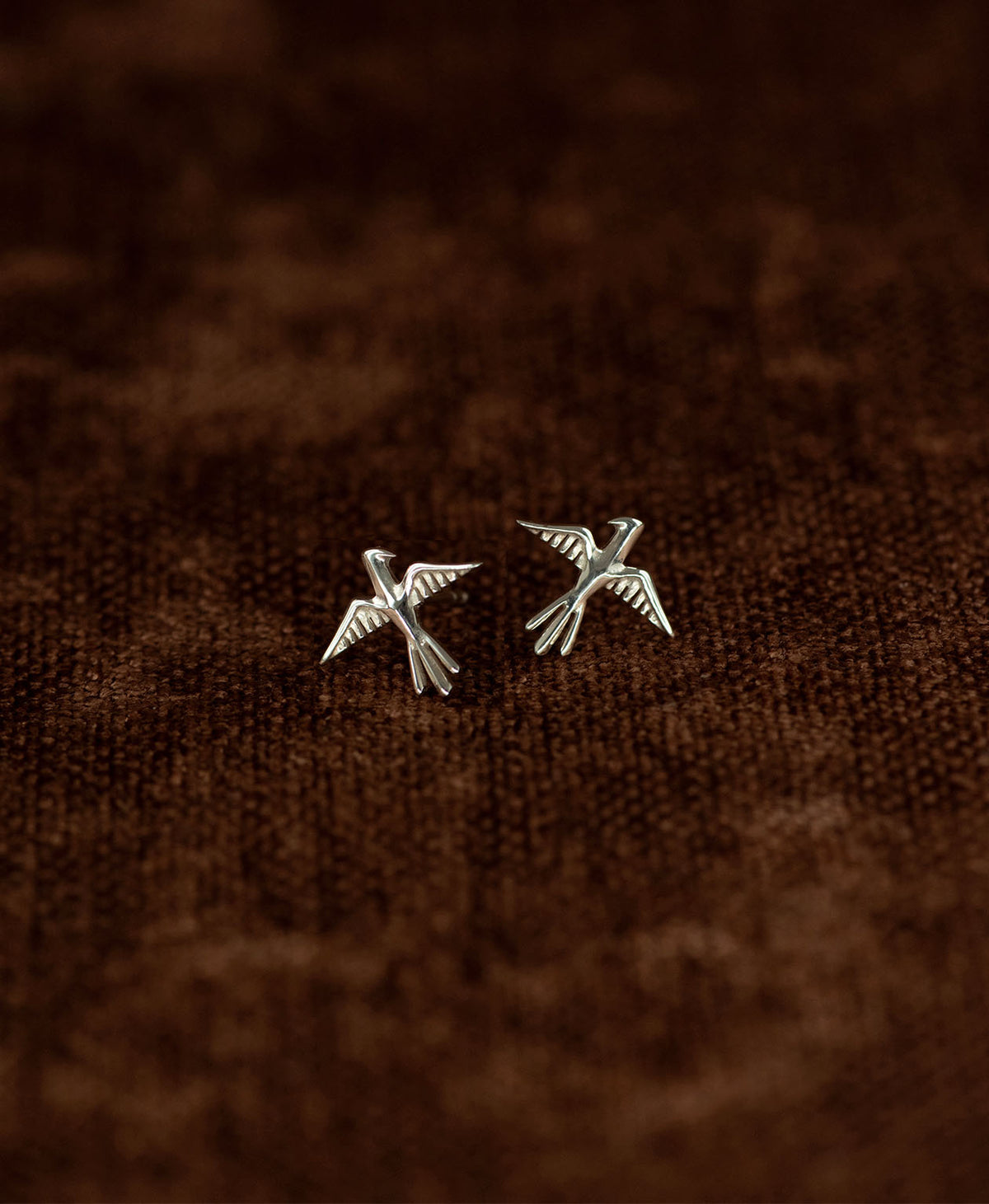 A pair of silver bird stud earrings with detailed feather and wing textures, displayed on a dark brown textured fabric background.
