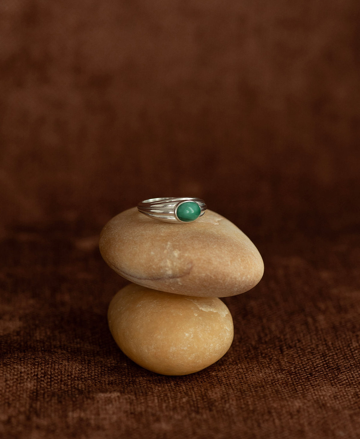 The silver Esmeralda Ring, featuring a cabochon cut Emerald set in a graduated band, rests on a cream stone on a textured brown background. 