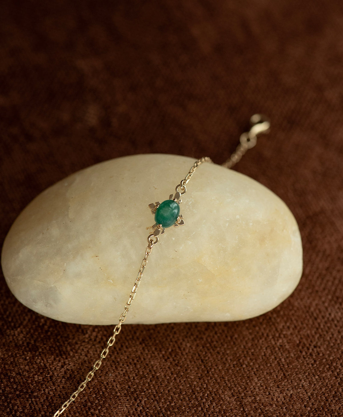 A close up of the gold Farah Bracelet featuring a cabochon cut emerald, layered over a cream stone on a textured brown fabric background.