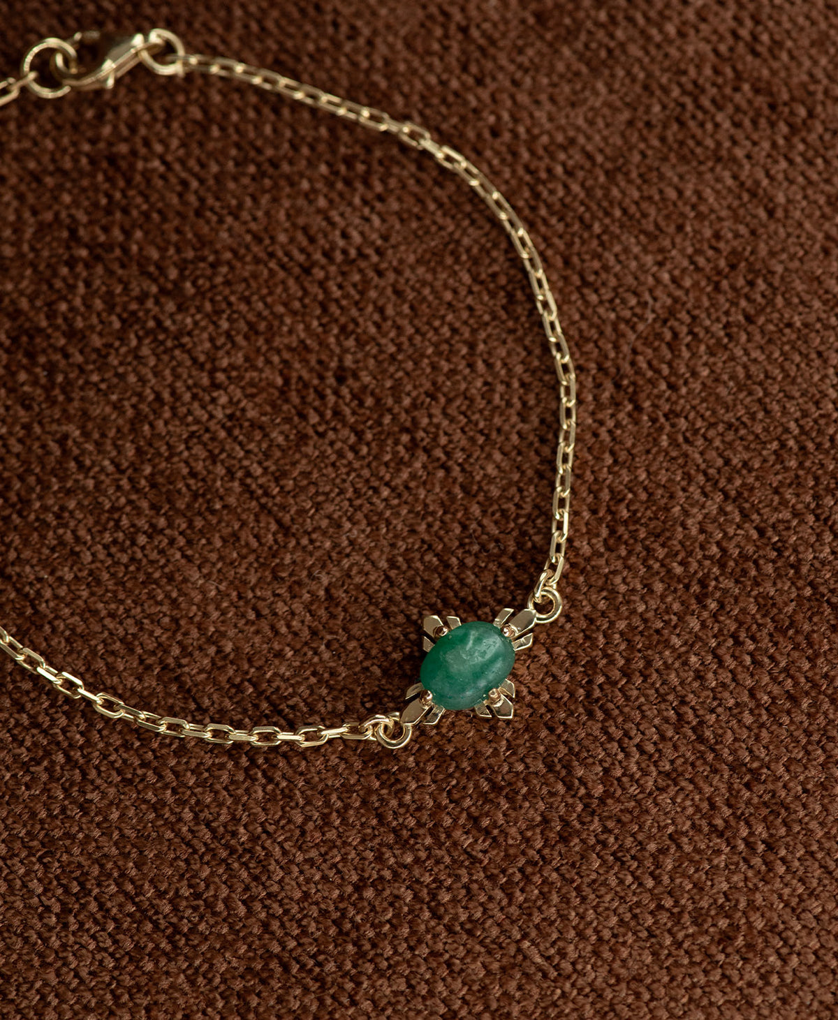 A close up of the gold Farah Bracelet featuring a cabochon cut emerald, on a textured brown fabric background.