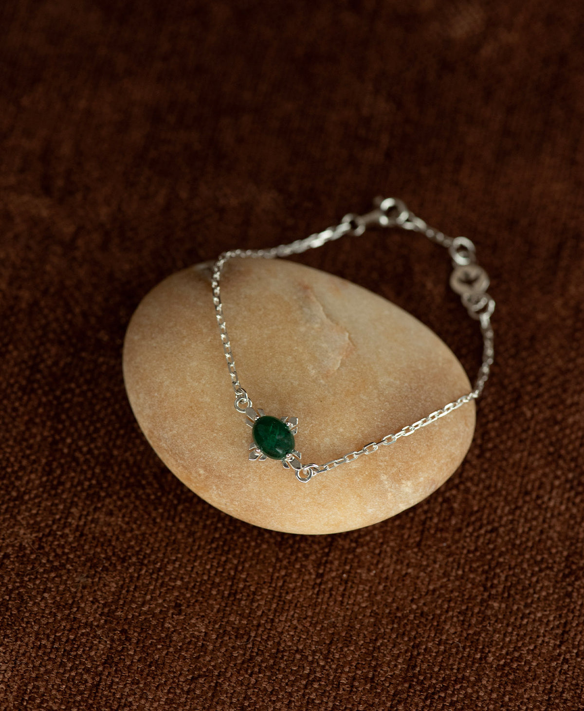 A close up of the silver Farah Bracelet featuring a cabochon cut emerald, layered over a cream stone on a textured brown fabric background.