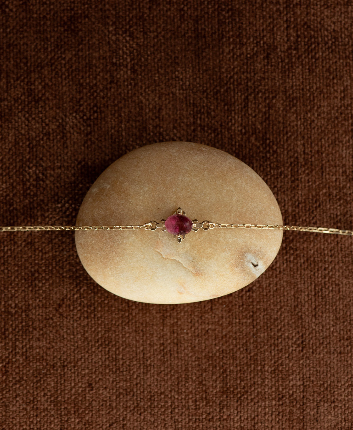 A close up of the gold Farah Bracelet featuring a cabochon cut pink tourmaline, layered over a cream stone on a textured brown fabric background.