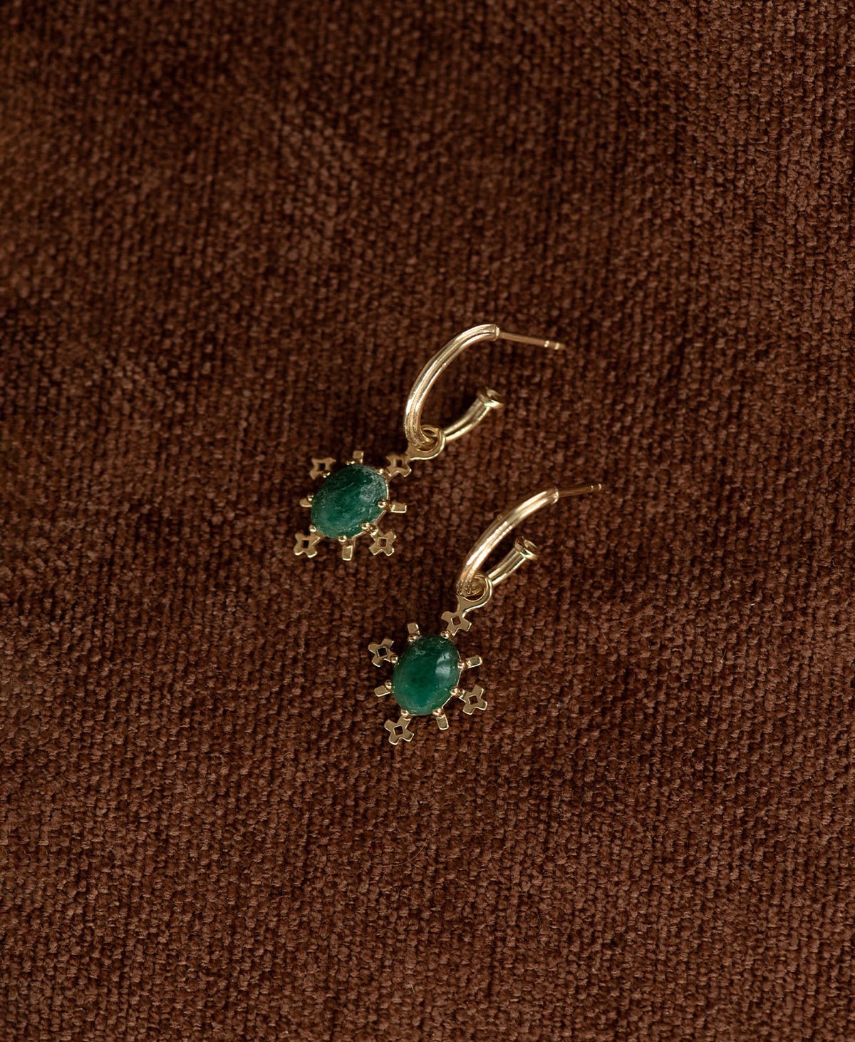 A pair of Farah gold drop earrings with emerald gemstones, displayed against a dark brown textured background.