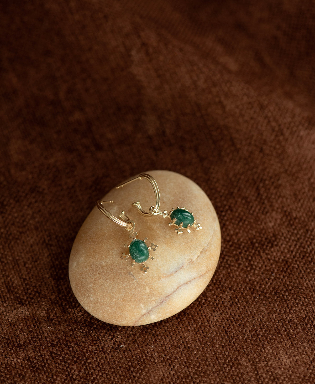 A pair of Farah gold drop earrings with emerald gemstones, displayed on a smooth cream coloured stone against a dark brown textured background.