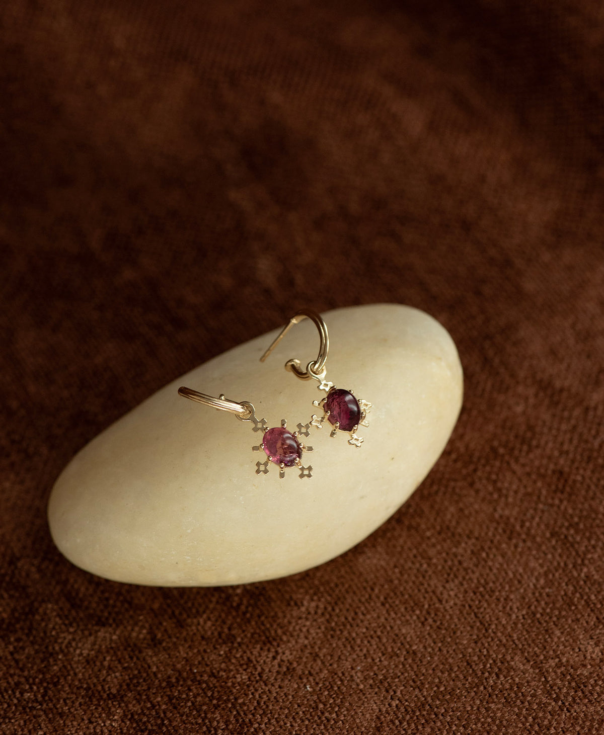 A pair of Farah gold drop earrings with pink tourmaline gemstones, displayed on a smooth cream coloured stone against a dark brown textured background.